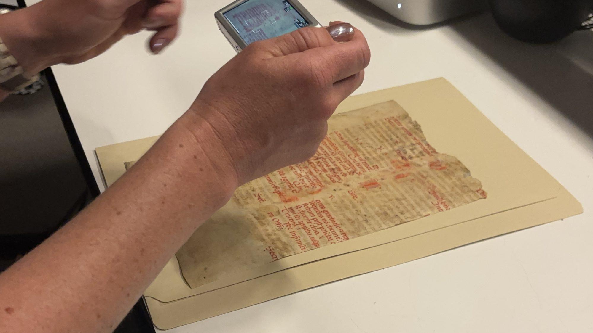 A manuscript leaf being photographed from above. Hands hold a small digital camera in frame.